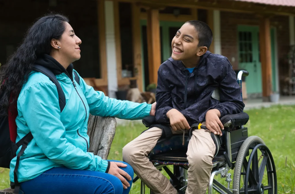 A woman sits on a bench outdoors, smiling at a boy in a wheelchair who is laughing. They are in front of a building with green doors and wooden beams.